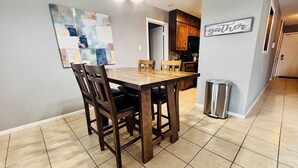 A dining area with a wooden table and chairs, a kitchen in the background, and a trash can.