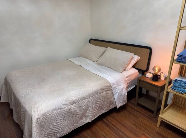 A neatly made bed with a light-colored quilt, a bedside table with a lamp, and a wooden shelf with folded items.