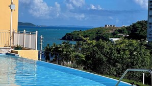 Una piscina con agua azul cristalina, un edificio amarillo y vistas al océano y colinas distantes.