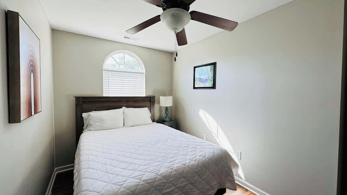 A bedroom with a bed, a ceiling fan, a window with blinds, a nightstand with a lamp, and a framed picture on the wall.