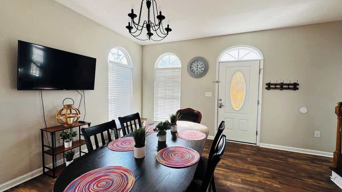 Un área de comedor con una mesa de madera oscura, sillas negras y posavasos circulares rosa. Un candelabro cuelga del techo y un televisor de pantalla plana está montado en la pared.