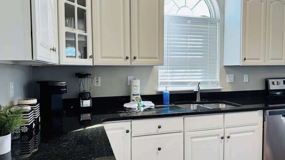 A kitchen with white cabinets, a black countertop, a coffee maker, a blender, and a window with blinds.