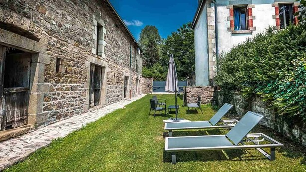 Un edificio de piedra con un patio cubierto de césped, dos sillones de descanso y una pequeña mesa con sombrilla.