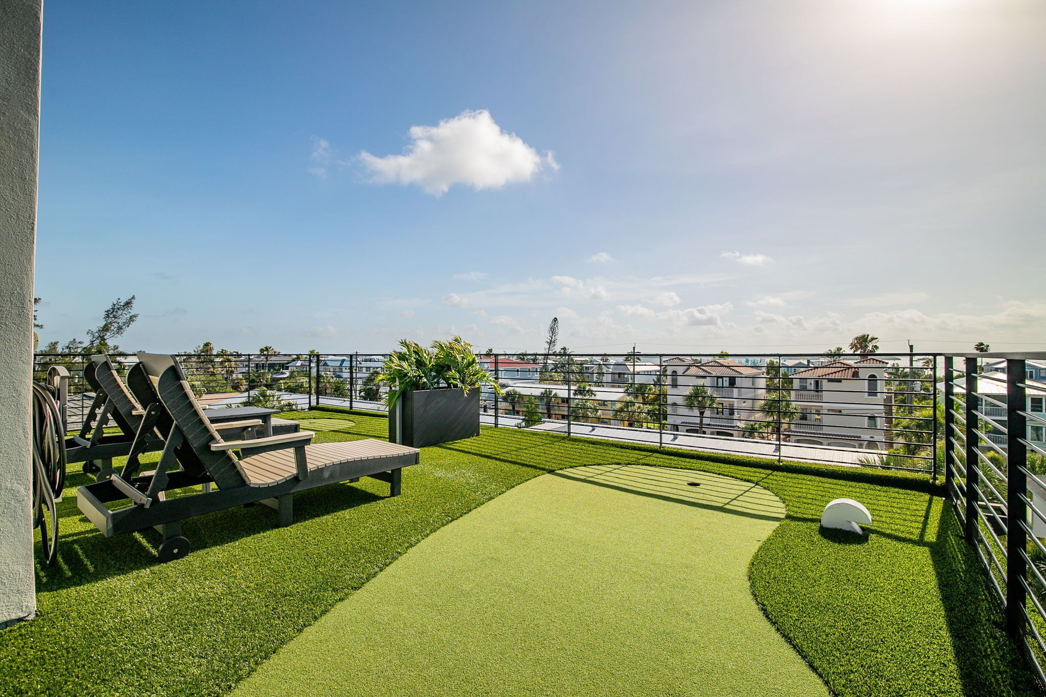 A rooftop terrace with artificial grass, lounge chairs, a planter with plants, and a view of buildings in the distance.