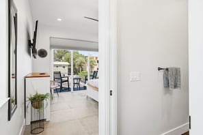 Un interior moderno con una pared blanca, un televisor montado, una planta en maceta y vistas a un balcón exterior con sillas y una mesa.