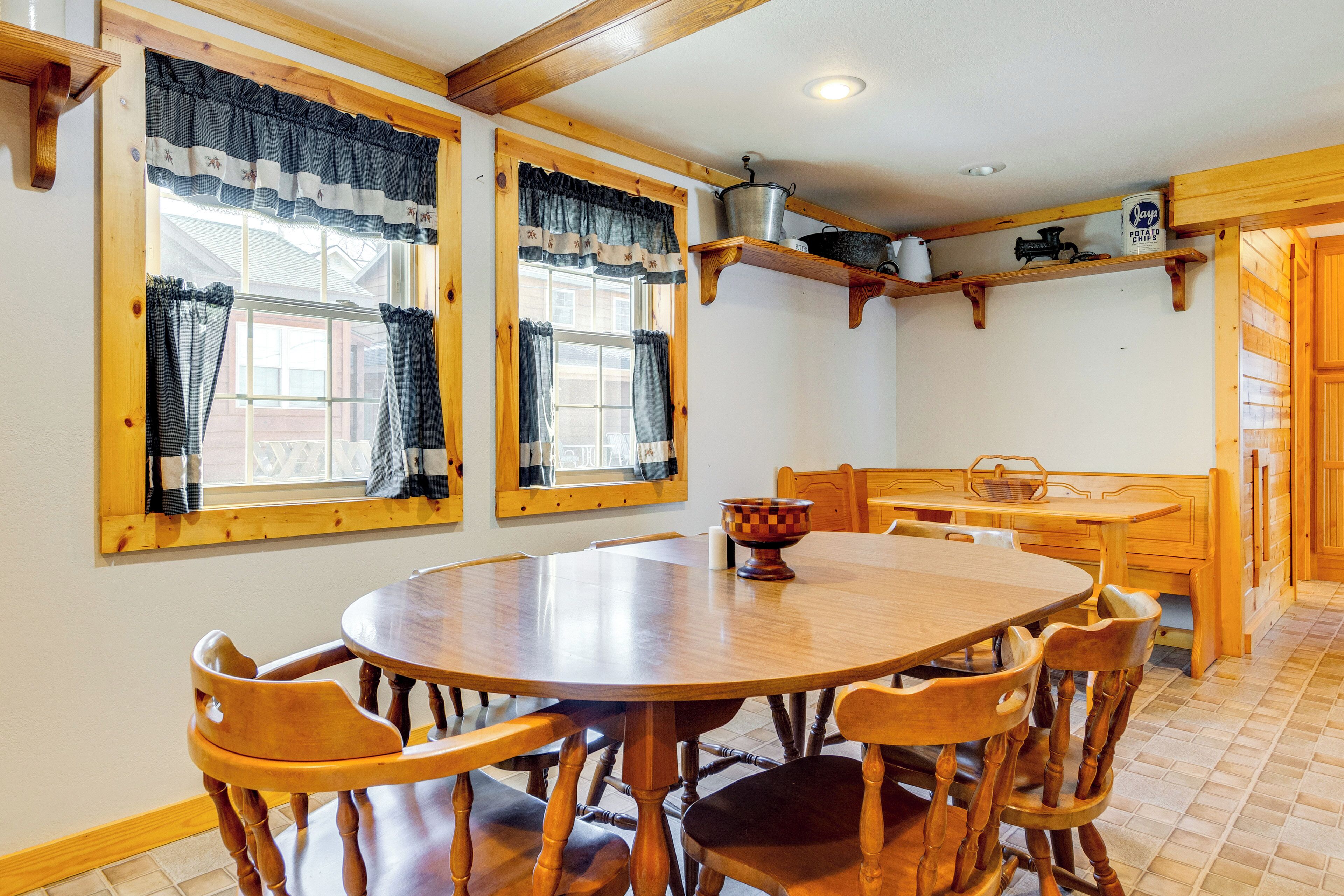 A wooden dining table with six chairs, a window with curtains, and a shelf with kitchen items.
