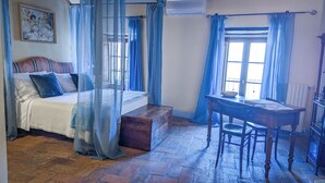 A bedroom with a four-poster bed, a wooden chest, a desk with a chair, and a window with blue curtains. - PienzaLettings Suites (Pienza)