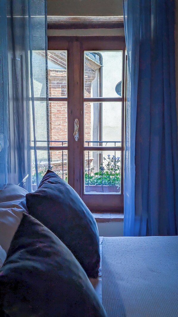 A room with a bed, a window with curtains, and a view of a brick building outside.