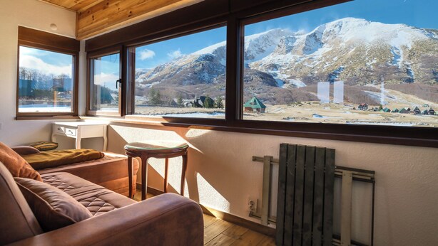 Une pièce chaleureuse avec un plafond en bois, de grandes fenêtres, un canapé en cuir marron, une petite table et une vue sur des montagnes enneigées.