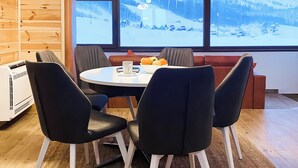 Une pièce chaleureuse avec un décor intérieur en bois, une table ronde entourée de chaises, une vue sur un paysage enneigé par de grandes fenêtres et un radiateur au sol.