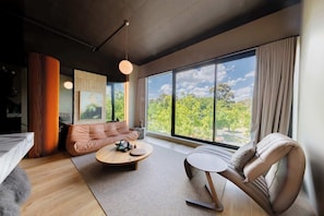 Een moderne kamer met een bank, een stoel, een bijzettafeltje en een groot raam met uitzicht op bomen.
