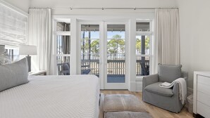 A bedroom with a bed, a grey armchair, a small table with a lamp, and a view of an outdoor balcony with a railing. - Elevated Vacation Living• Fairmont Inspiration by the Bay (Miramar Beach)
