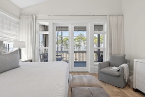 A bedroom with a bed, a grey armchair, a small table with a lamp, and a view of an outdoor balcony with a railing.