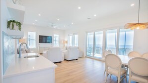 A modern kitchen with a white countertop and sink, a dining area with a white table and chairs, and large windows offering a view of the city. - Gulf front, elevator, private pool, outdoor dining, beach gear! (PORT SAINT JOE)