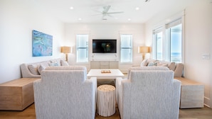 A modern living room with a white ceiling fan, a flat-screen TV, and a large window with blinds. - Gulf front, elevator, private pool, outdoor dining, beach gear! (PORT SAINT JOE)