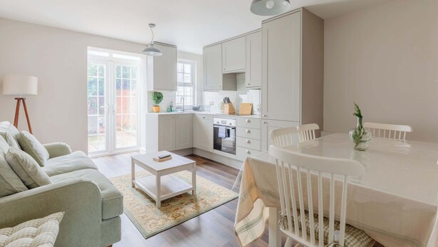 A modern kitchen with white cabinets, a dining table, and a sofa in a bright living area with a large window. - Cosy 4BD Flat in Stockwell with Private Patio (Greater London)