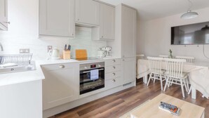 A modern kitchen with white cabinetry, a built-in oven, a dining area with white chairs, and a flat-screen TV mounted on the wall. - Cosy 4BD Flat in Stockwell with Private Patio (Greater London)