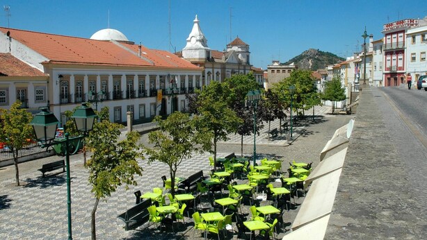 Una calle empedrada con mesas y sillas verdes al aire libre y edificios blancos con tejados de tejas rojas.