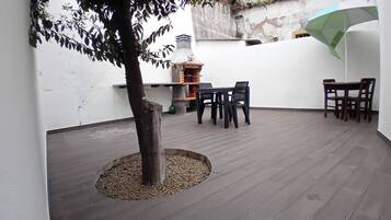 Un patio con mesa y sillas de madera, un pequeño árbol y un horno de ladrillo.