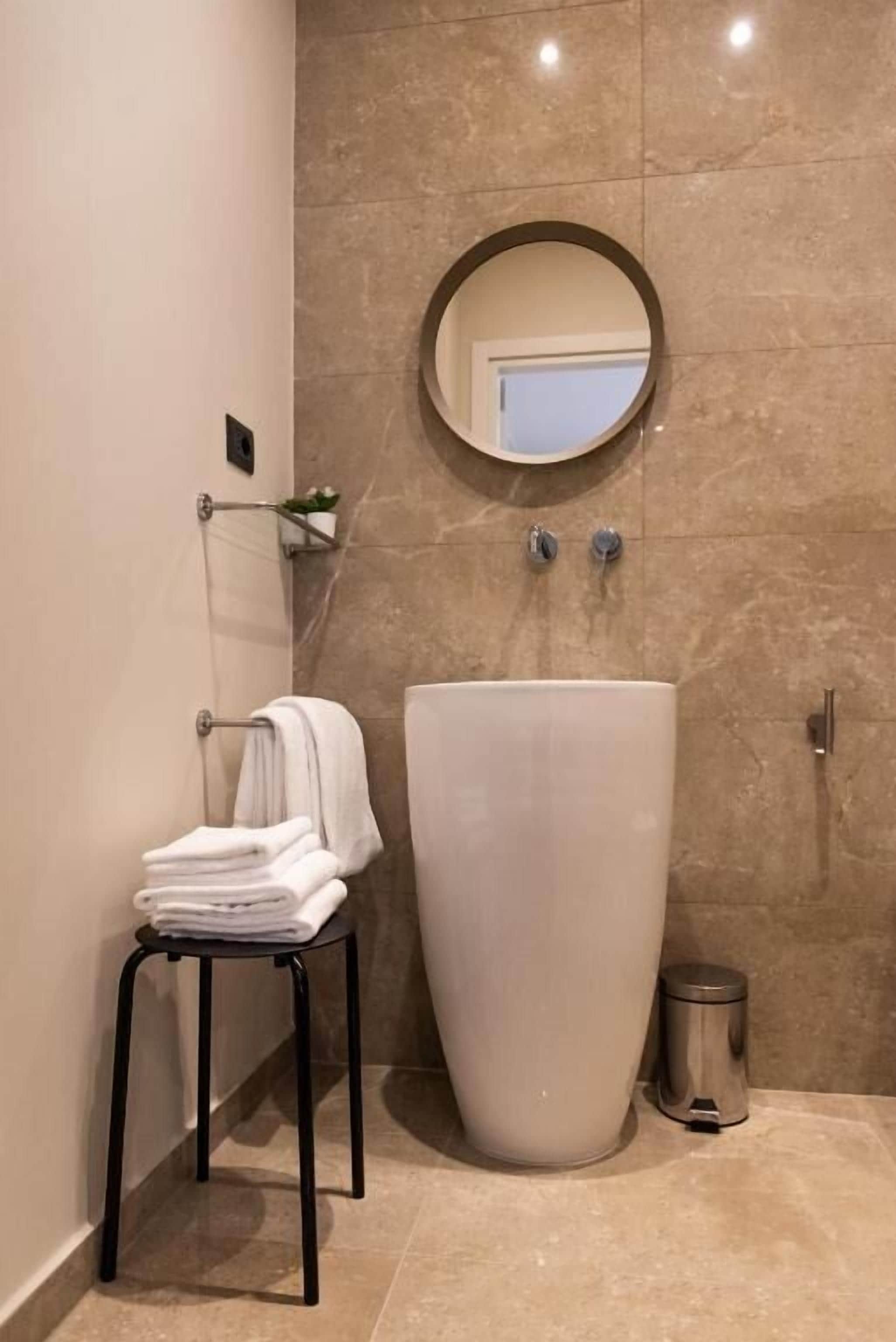 A bathroom with a large white sink, a round mirror, a towel rack with folded towels, and a trash can.