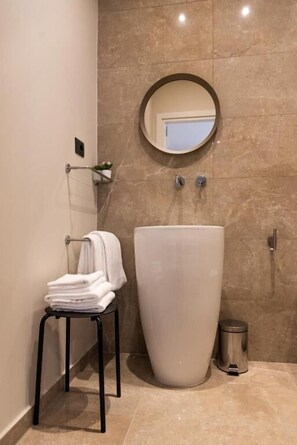 Un baño con un lavamanos grande de color blanco, un espejo redondo, un perchero con toallas dobladas y un cubo de basura.