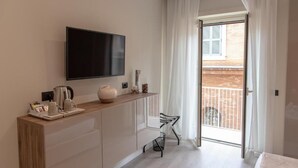 A modern hotel room with a flat-screen TV, a wooden console table with a kettle and cups, a chair, and a balcony with a view of a brick building. - XXIV Domus Luxury Suites (Rome)