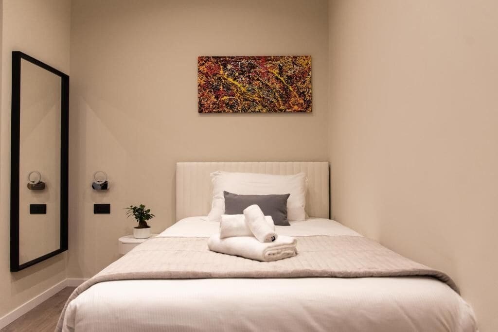 A hotel room with a bed, a painting on the wall, a small table with a plant, and a mirror on the wall.