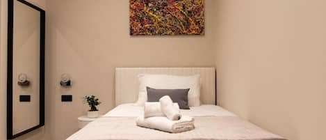 A hotel room with a bed, a painting on the wall, a small table with a plant, and a mirror on the wall.