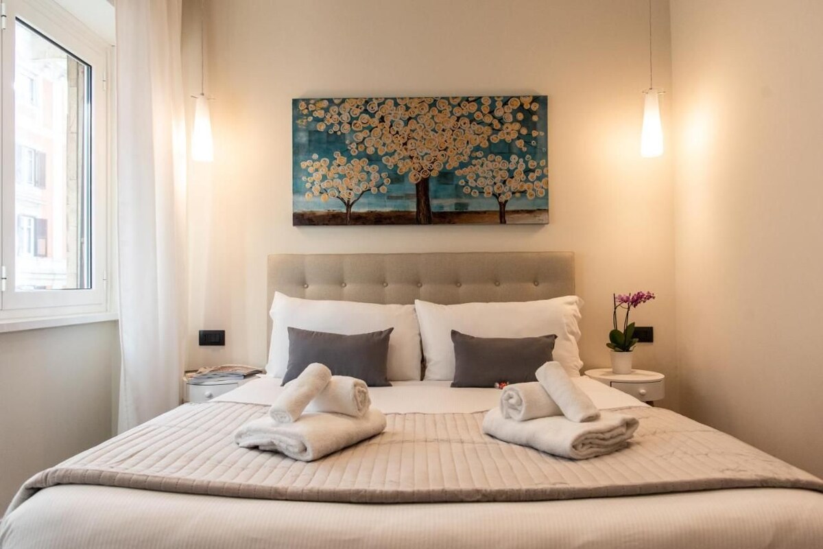 Un lit bien fait, avec deux oreillers, une table de chevet sur laquelle se trouve une lampe, et un tableau représentant des arbres accroché au mur.