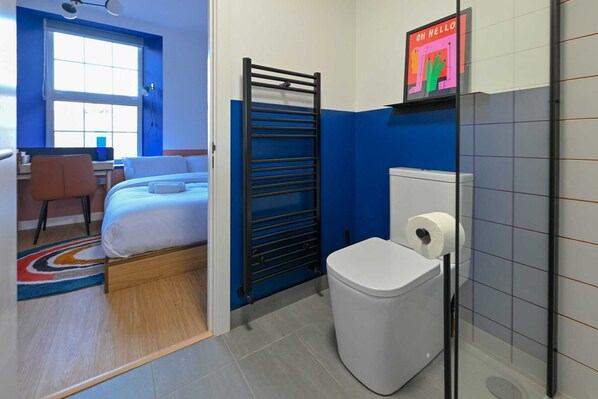 A hotel room with a bed, a desk, a chair, a radiator, a toilet, and a window.