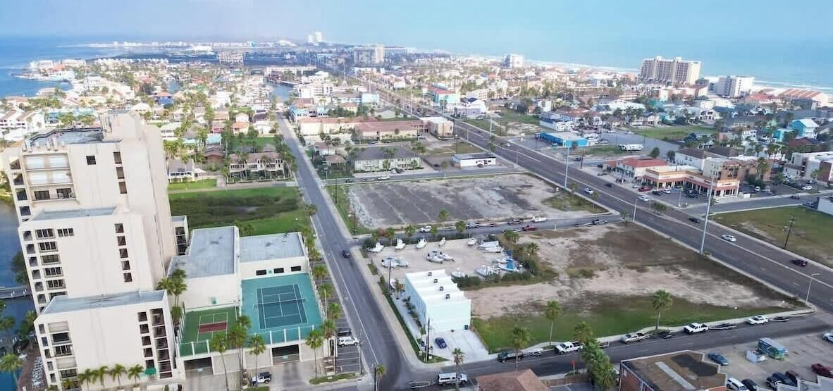 Vista aérea de una ciudad costera con una mezcla de edificios residenciales y comerciales, un edificio blanco destacado con techo verde, y una vista despejada del océano.