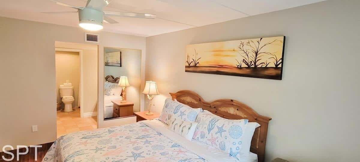 A bedroom with a bed, bedside tables, a nightstand, a ceiling fan, and a painting on the wall.