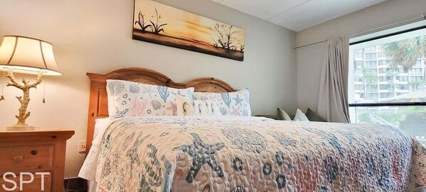 Un dormitorio con cama, una mesita de noche con lámpara, un cuadro en la pared y una ventana con cortinas.