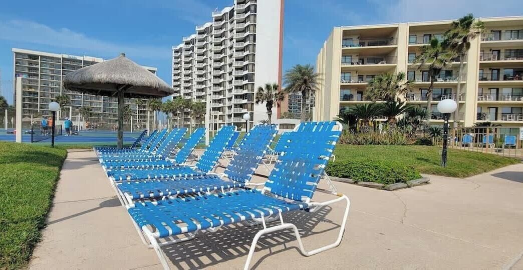 A poolside area with blue lounge chairs, a thatched umbrella, and a high-rise hotel building in the background.