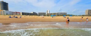 Una playa con edificios al fondo, una orilla arenosa y olas.
