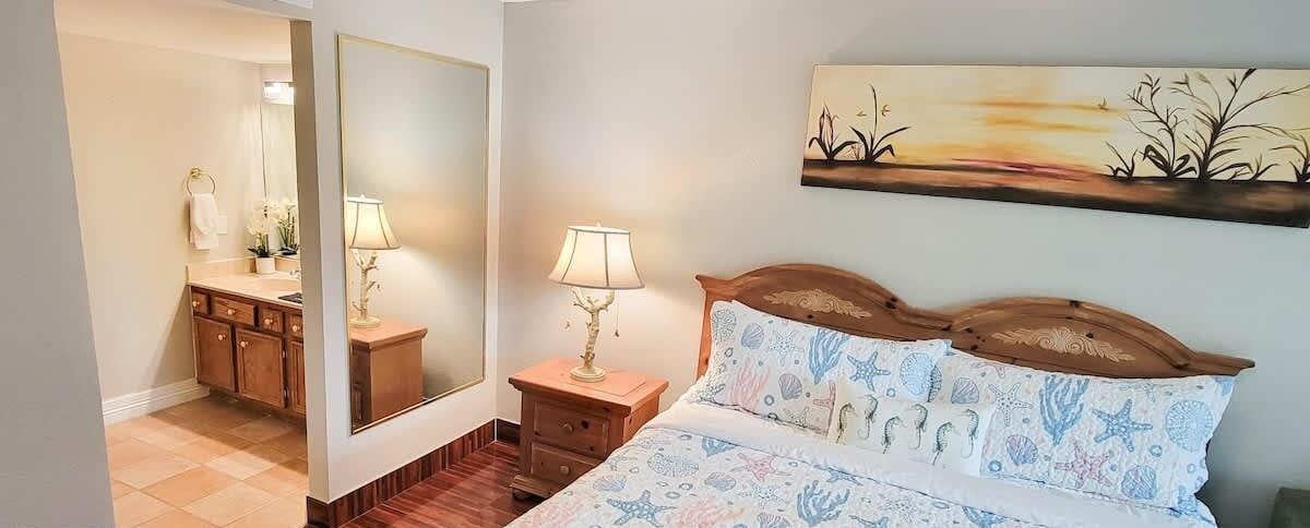 Un dormitorio con cama, mesitas de noche, un espejo, un cuadro y un baño visible a través de una puerta abierta.