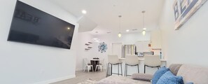 A modern living room with a flat-screen TV, a sofa with patterned cushions, a dining area with chairs, and a kitchen in the background. - Villas De Laguna 4 3 Bedroom Townhouse (South Padre Island)