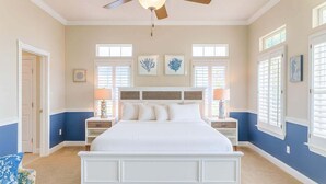 A bedroom with a large bed, two nightstands with lamps, a ceiling fan, and artwork on the walls.