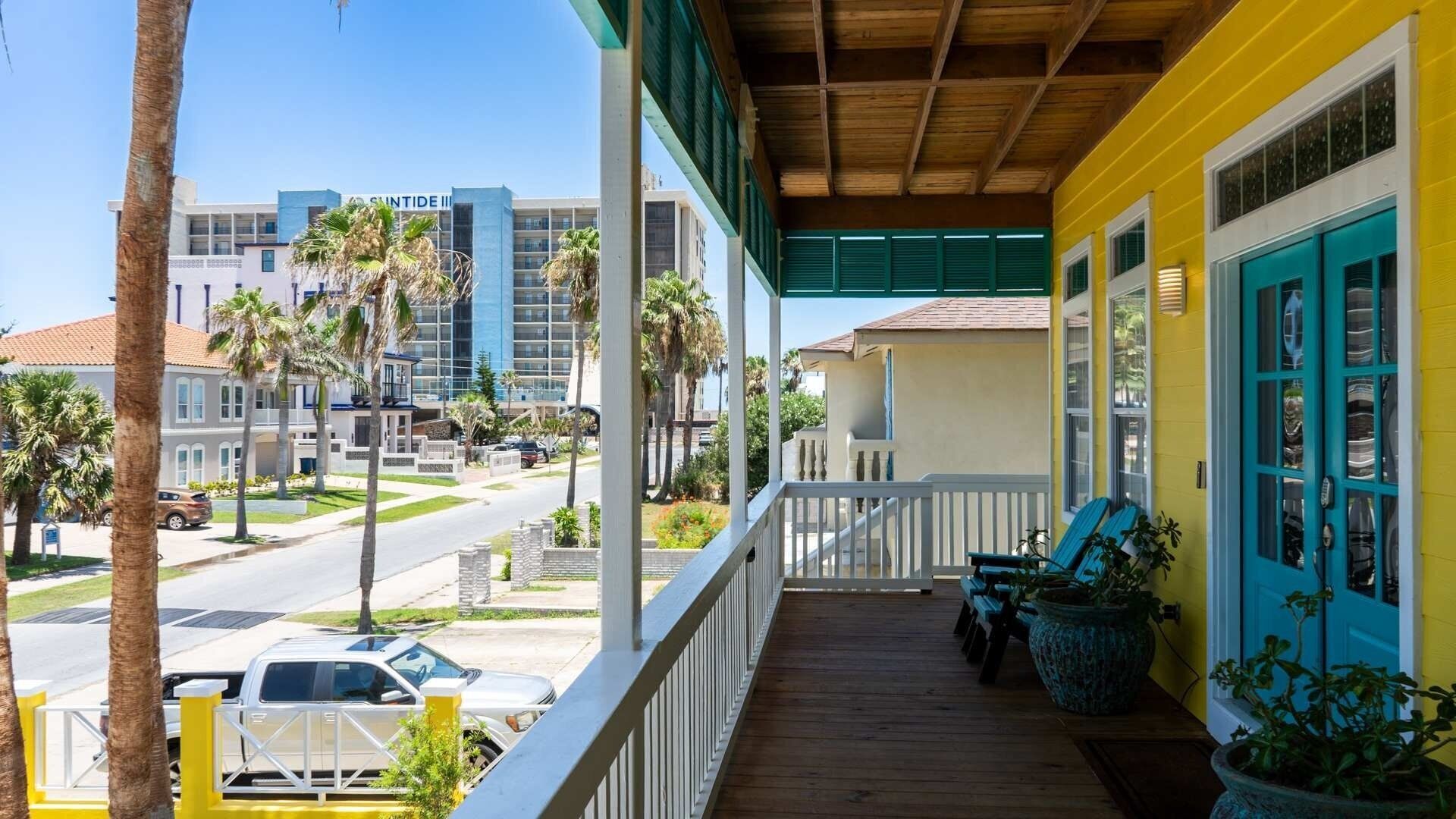 Un hôtel doté d’une terrasse en bois, d’une façade jaune et offrant une vue sur une rue bordée de palmiers et d’autres bâtiments.