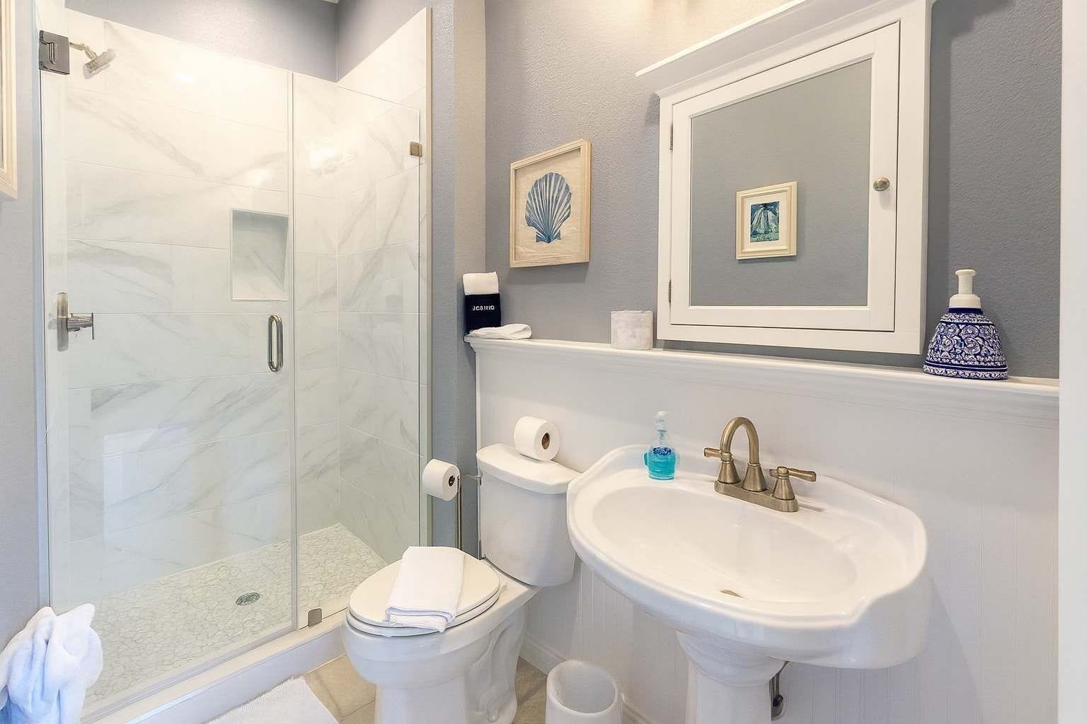 A bathroom with a glass shower enclosure, a toilet, a sink with a mirror above, and a blue decorative item on the sink.