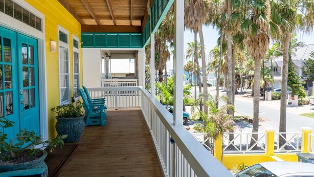 A wooden porch with teal chairs and potted plants, overlooking a street with palm trees and parked cars.