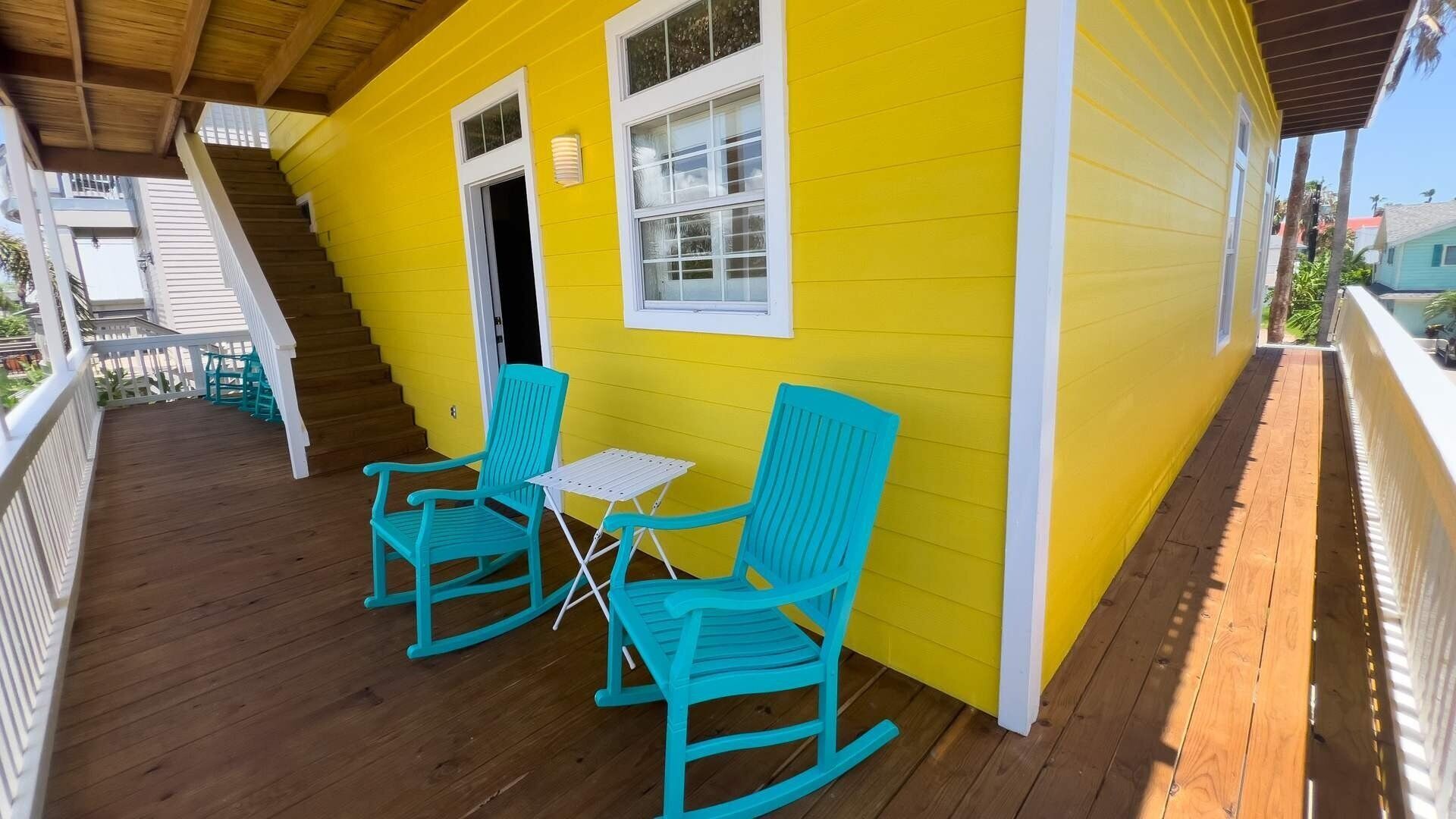 A yellow house with a wooden porch, two rocking chairs, and a small table.