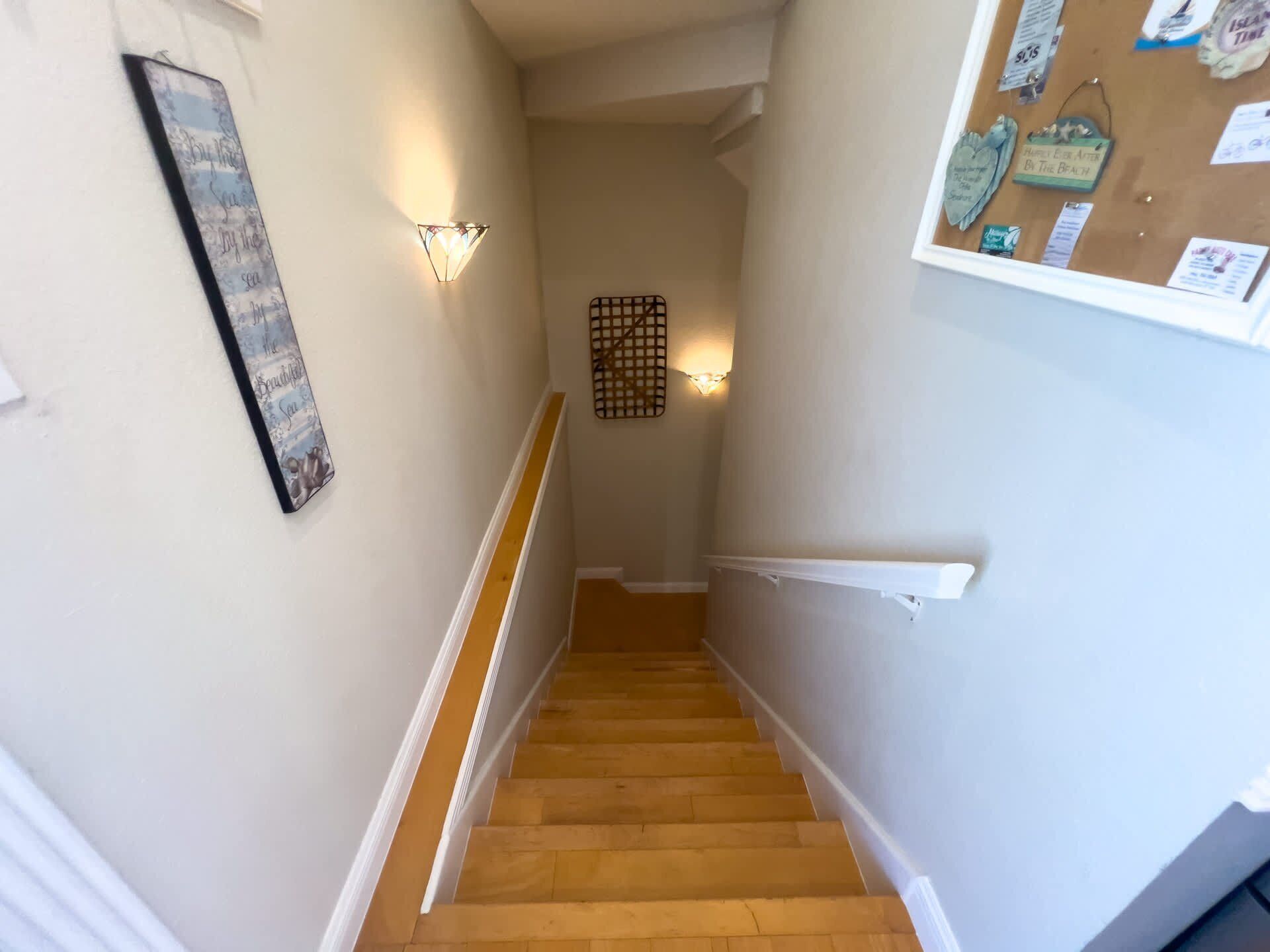 Una escalera con peldaños de madera y barandillas blancas que conduce a un rellano con un tablón de anuncios y luminarias fijadas a la pared.