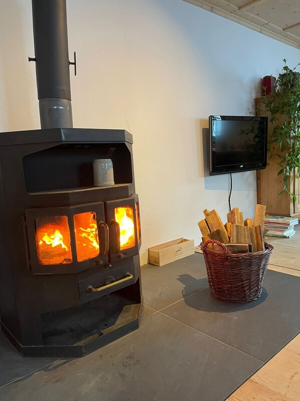 Un poêle à bois dont on peut voir les flammes, un téléviseur fixé au mur et un panier de bois de chauffage posé sur le sol.