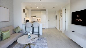 A modern living room with a sofa, a round coffee table, and two bar stools. There is a kitchen area with white cabinets and a dining table. A flat-screen TV is mounted on the wall.