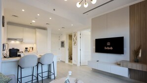 A modern living room with a kitchen area, a dining space, and a TV mounted on the wall. - Livbnb - Homey 1B with Spacious Terrace in JVC (Dubai)