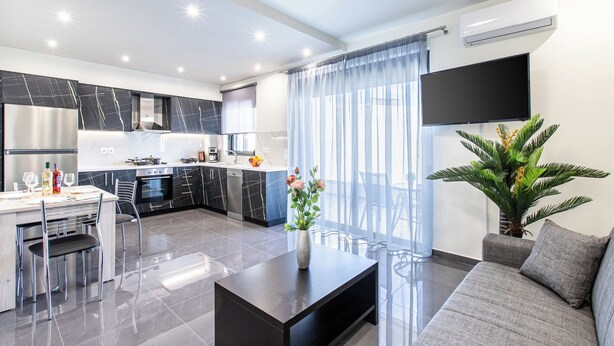 A modern kitchen with a dining area, a flat-screen TV, and a living room with a sofa and a plant.