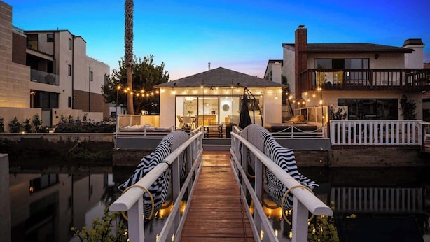 A waterfront property with a wooden deck leading to a glass-enclosed patio area, surrounded by modern buildings and a clear sky.
