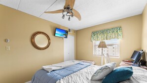 Ein Schlafzimmer mit einem Bett, einem Deckenventilator, einem Fernseher, einem Fenster mit Vorhängen und einem Schreibtisch mit Computer.