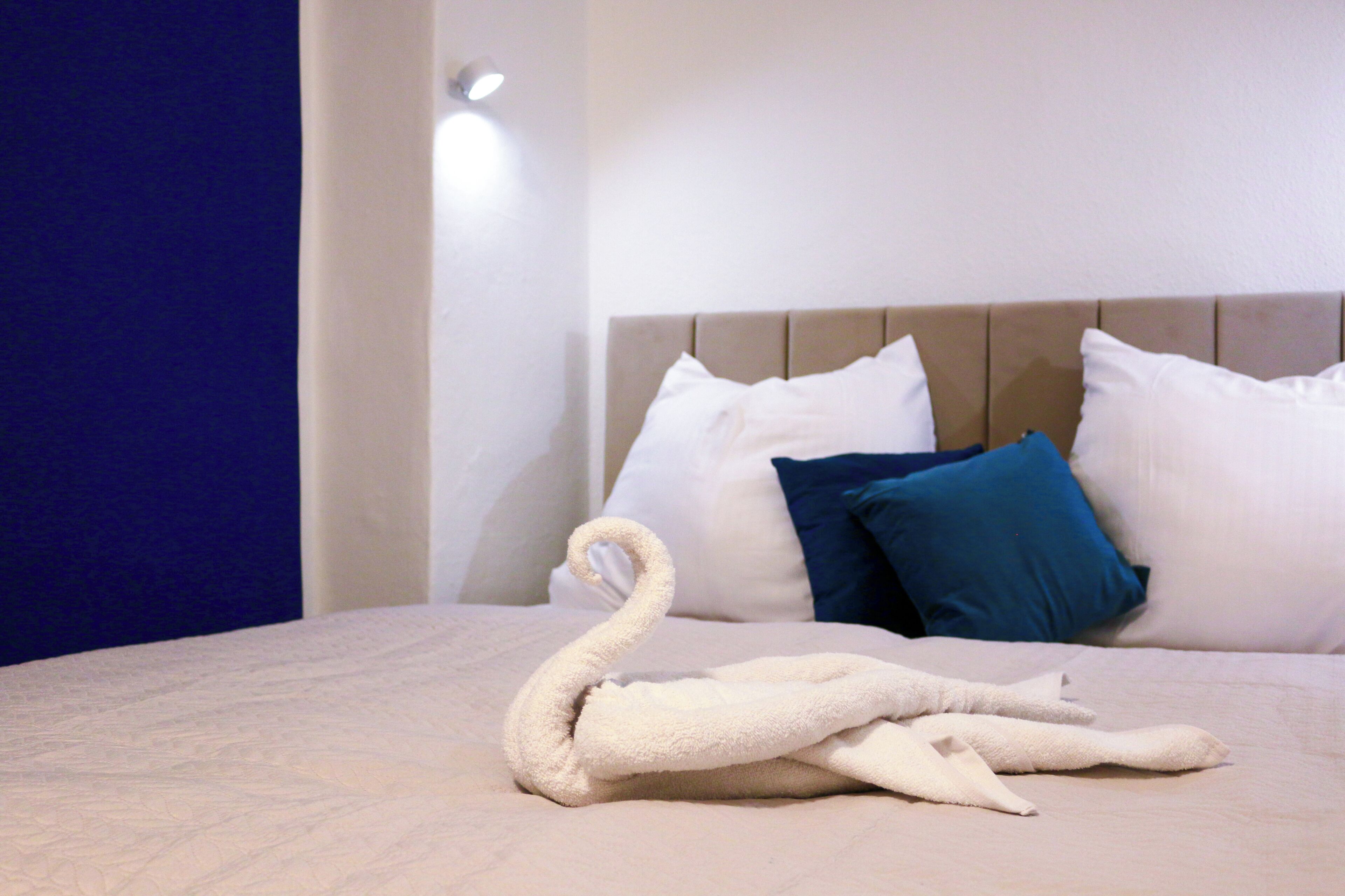 Een netjes opgemaakt bed met een handdoek in de vorm van een zwaan en twee kussens, één wit en één blauw, in een kamer met een effen muur en een lamp.